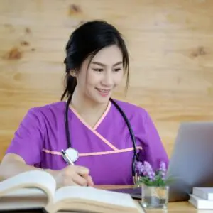 A woman wearing a violet uniform with a stethoscope on her neck sitting in front of a laptop