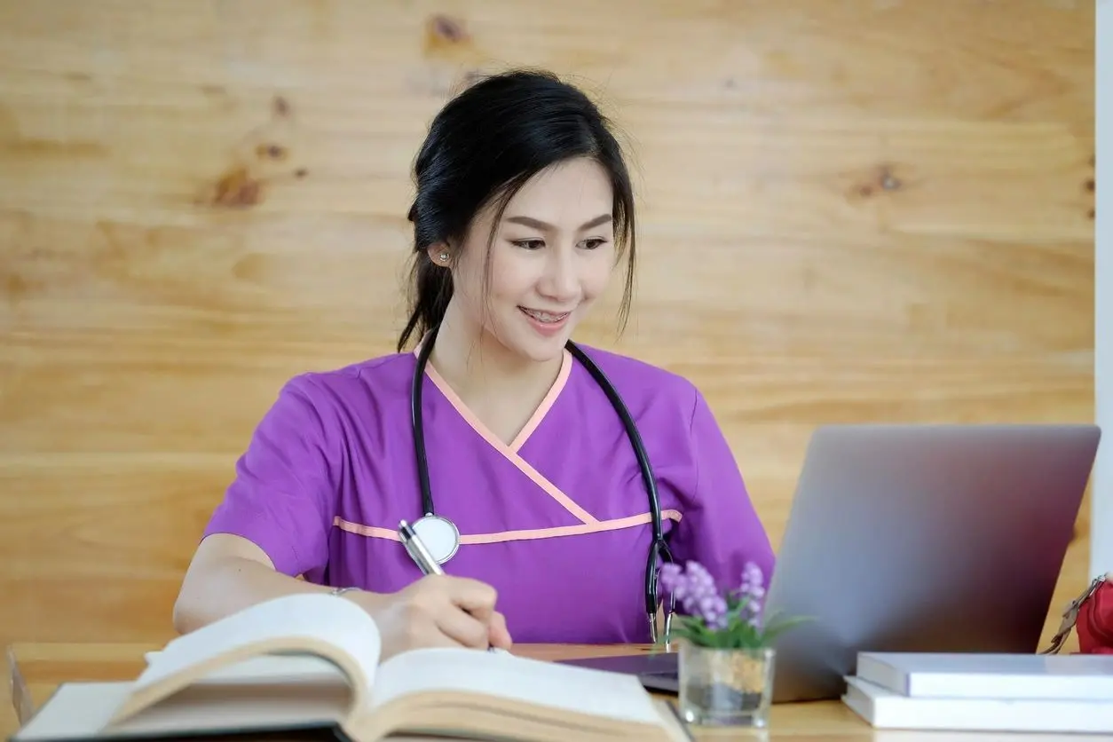 A woman wearing a violet uniform with a stethoscope on her neck sitting in front of a laptop